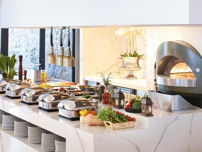 Buffet mit warmen Speisen, frischem Gemüse und Pizzaofen in modernem, hellem Raum.