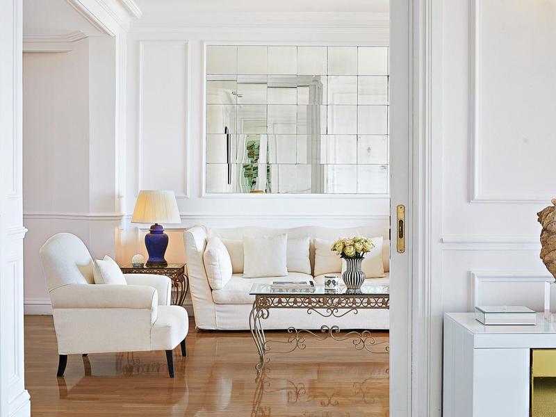 Helles Wohnzimmer mit weißen Möbeln, Glas-Couchtisch, blauer Vase und großem rechteckigem Spiegel.