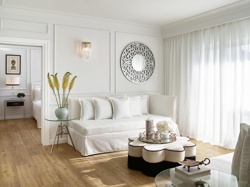 Helles Wohnzimmer mit weißem Sofa, runder Spiegel, Glastisch und Holzboden, große Fenster mit Vorhängen.