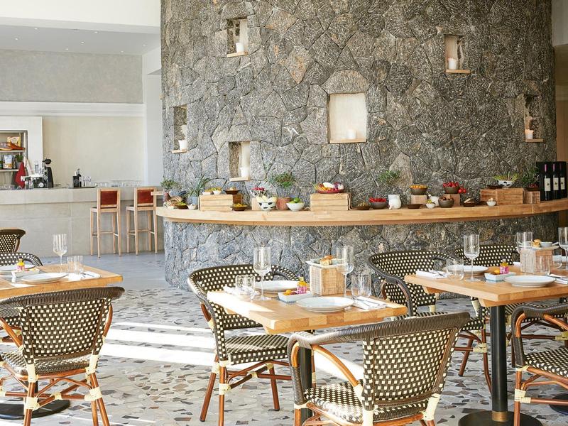 Helles Café mit runden Tischen, geflochtenen Stühlen und rustikaler Steinwand im Hintergrund.