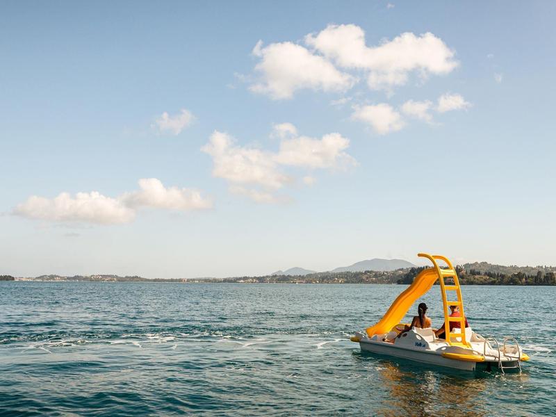 Tretboot mit gelber Rutsche auf ruhigem See unter blauem Himmel mit wenigen weißen Wolken.