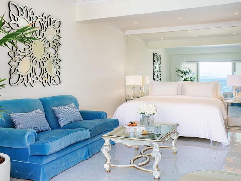 Wohnzimmer und Schlafzimmer kombiniert, blaue Couch, weißes Bett, Glas-Couchtisch, helle Wände.