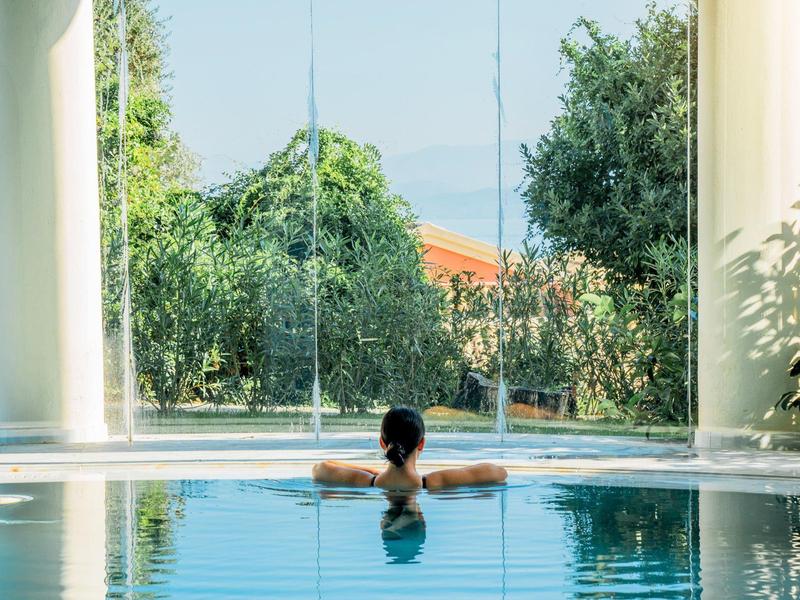 Frau entspannt im Pool, umgeben von großen Fenstern mit Blick auf grüne Bäume und blauem Himmel.