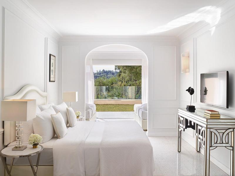 Helles Schlafzimmer mit großem Bett, weißen Wänden, TV auf Kommode und Blick auf Garten durch Bogenfenster.