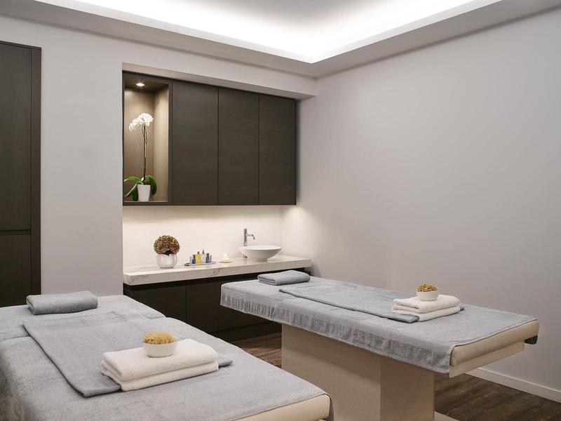 Behandlungsraum mit zwei Liegen, grauen Bezügen, Handtüchern und gedämpftem Deckenlicht.