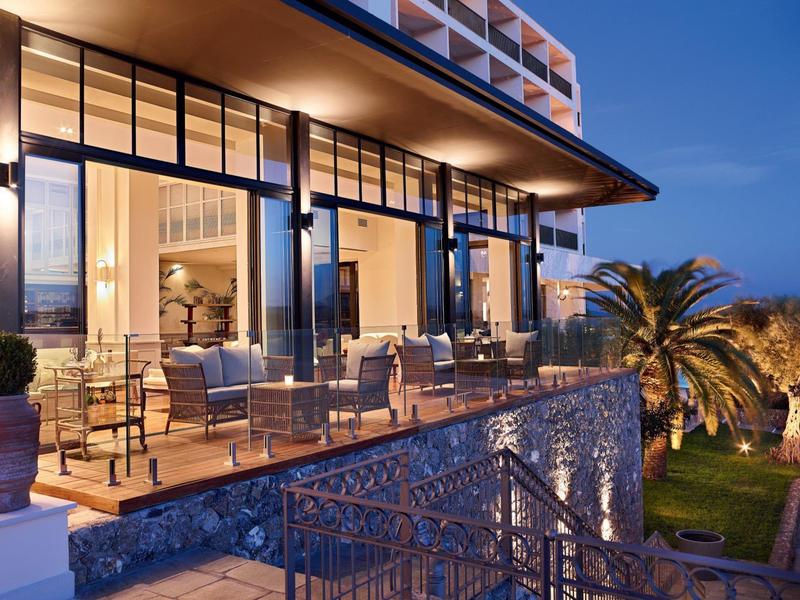 Moderner Balkon mit Glasgeländer, Stühlen und Tischen, beleuchtet bei Abenddämmerung.