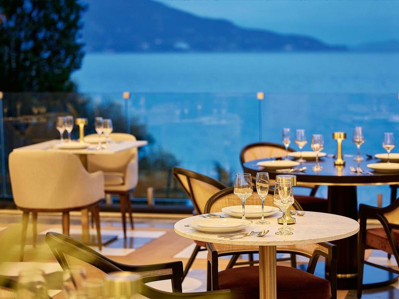 Elegantes Restaurant mit runden Tischen, Stühlen und Glaswand mit Meer- und Bergblick bei Abendlicht.