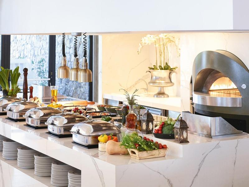 Buffet mit warmen Speisen, frischem Gemüse und Pizzaofen in modernem, hellem Raum.