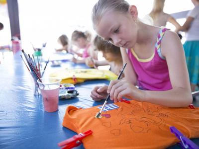 Mehrere Kinder gestalten bunte T-Shirts an einem langen Tisch bei einem Freizeitangebot.