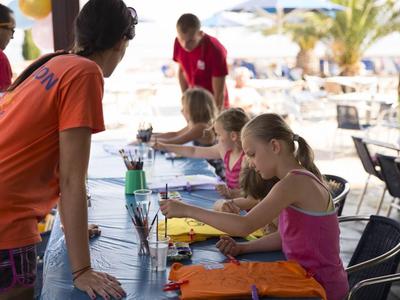 Kinder sitzen an einem Tisch im Freien und basteln unter Aufsicht eines Betreuers.