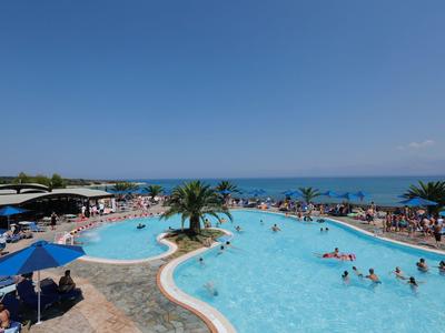 Großer Hotelpool mit Palmen und Sonnenschirmen am Meer unter klarem blauem Himmel.