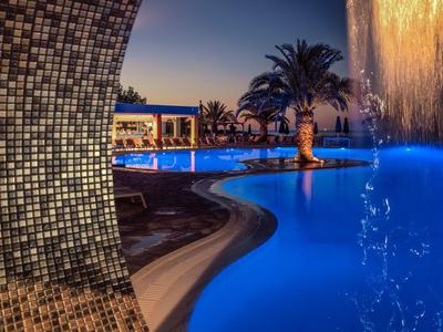 Beleuchteter Poolbereich mit Wasserfall und Palmen in einem Hotel bei Nacht.