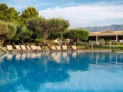 Großer Pool vor Sonnenliegen und Bäumen in einer Hotelanlage unter blauem Himmel.