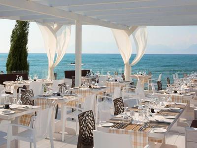 Ansicht eines offenen Restaurants mit weißen Tischen und Stühlen am Meer unter einem weißen Pavillon.
