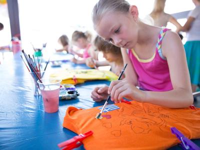 Kinder malen konzentriert auf orangefarbene Stofftaschen an einem hellen Basteltisch.