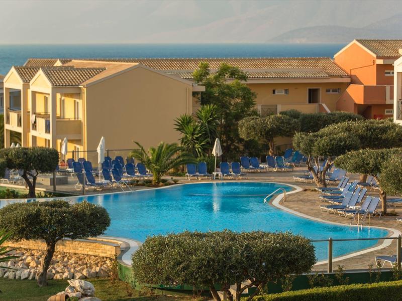 Ein blauer Swimmingpool mit Liegestühlen umgeben von Büschen und Gebäuden am Meer im Hintergrund.