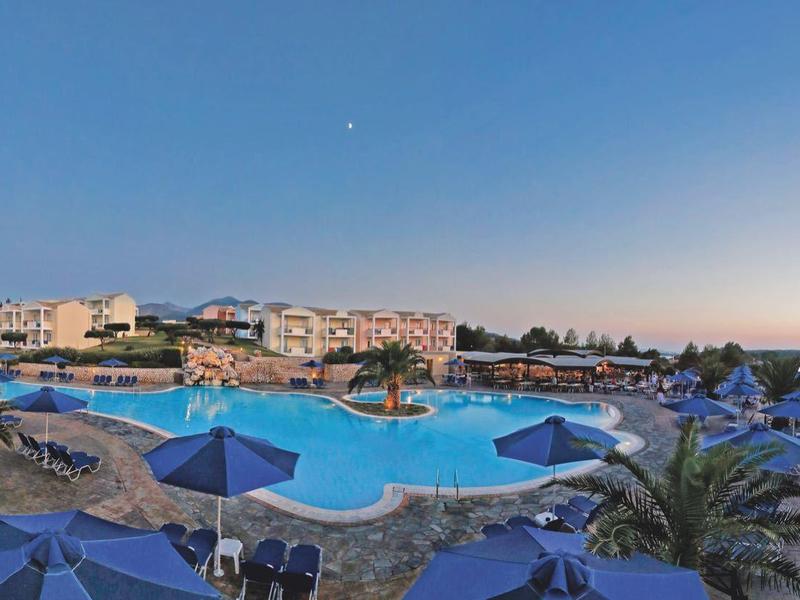 Großer blauer Pool mit Sonnenschirmen und Palmen an Küste bei klarem Himmel und Sonnenuntergang.