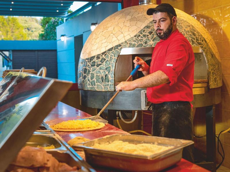 Mann in rotem Hemd belegt Pizza vor einem großen Holzofen in einer farbenfrohen Pizzeria.