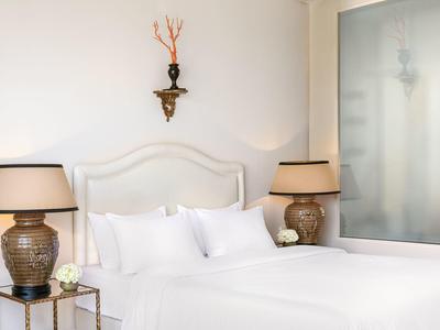 Weißes Bett mit Kopfteil, zwei Tischlampen und Wanddeko in einem hellen Hotelzimmer.