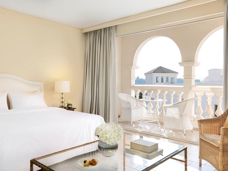 Helles Hotelzimmer mit großem Bett, Tisch, Stühlen und Blick auf eine mediterrane Stadt.