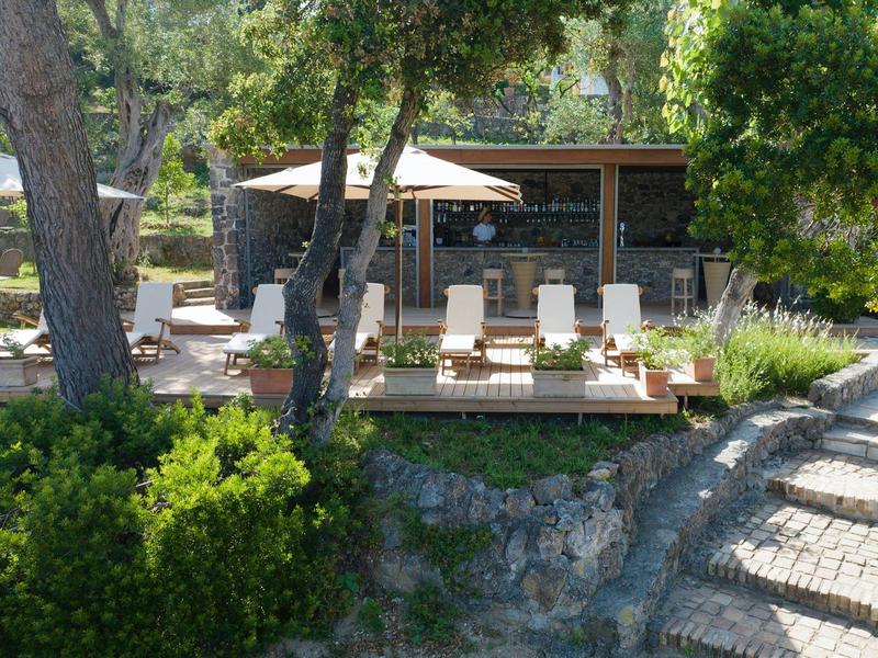 Terrasse mit Liegestühlen und Sonnenschirmen vor einem Steinhaus, umgeben von Bäumen und Pflanzen