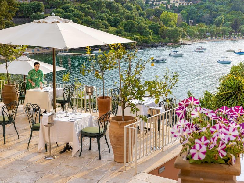 Terrasse mit Tischen, Stühlen und Sonnenschirm mit Blick auf einen bewaldeten See mit Booten.