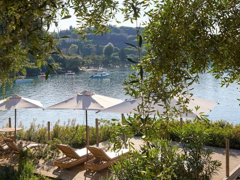 Blick auf einen See mit Sonnenschirmen und Liegestühlen, umgeben von Bäumen.