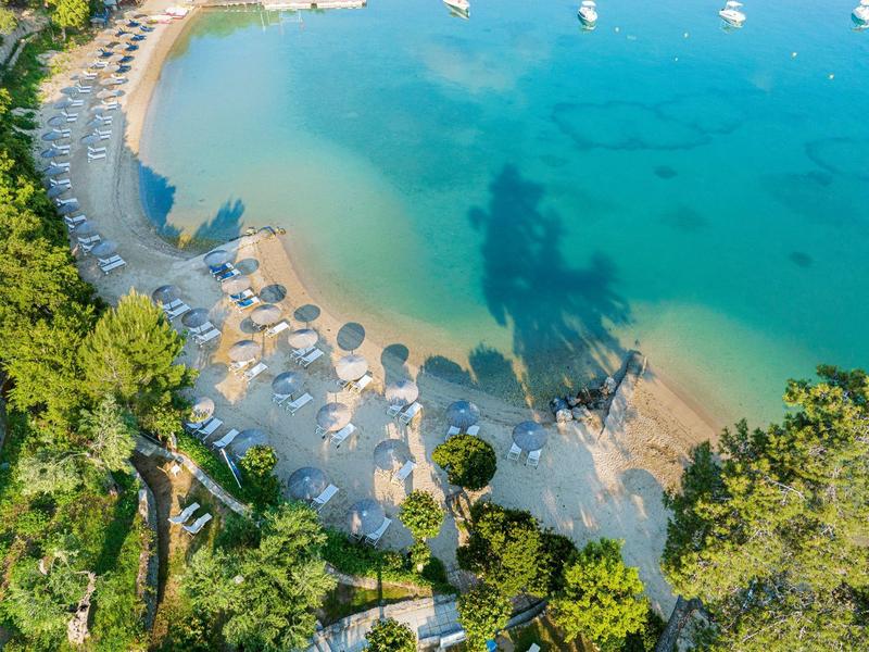 Sandstrand mit Sonnenschirmen und Liegen neben klarem, türkisblauem Wasser und grünen Bäumen.