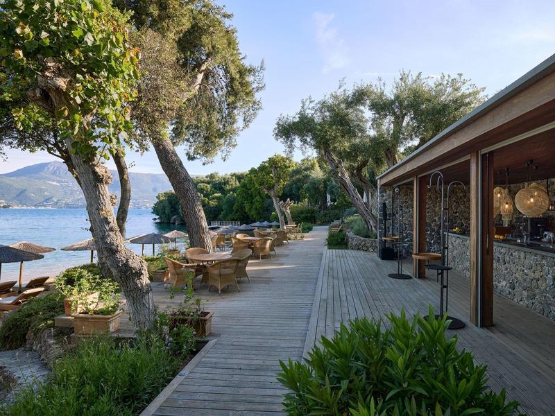 Holzterrasse mit Sitzgelegenheiten neben einem Hotel mit Blick auf das Meer und Bäume.