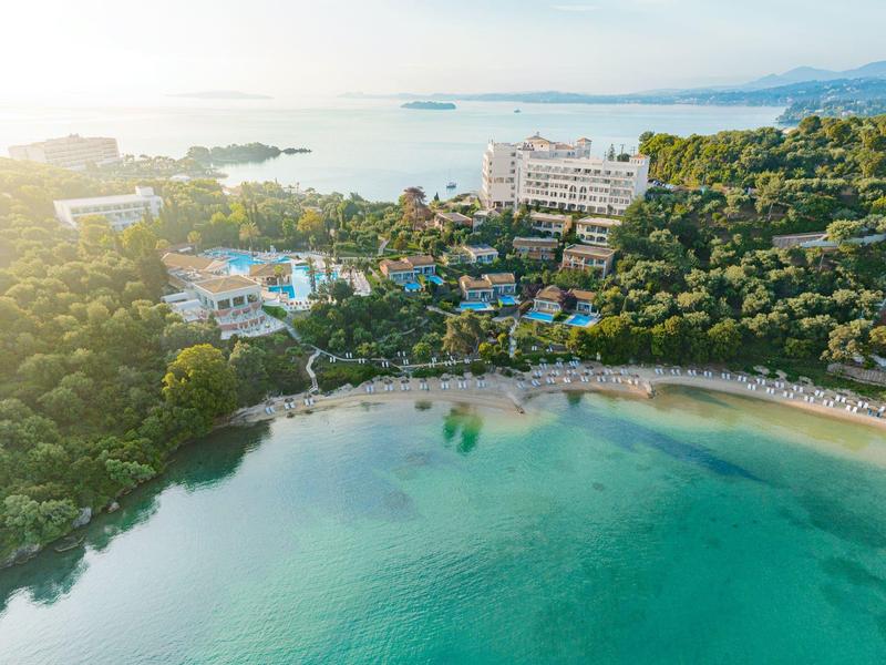 Luftaufnahme eines Strandes mit klarem Wasser, umgeben von Grünflächen und Hotels.