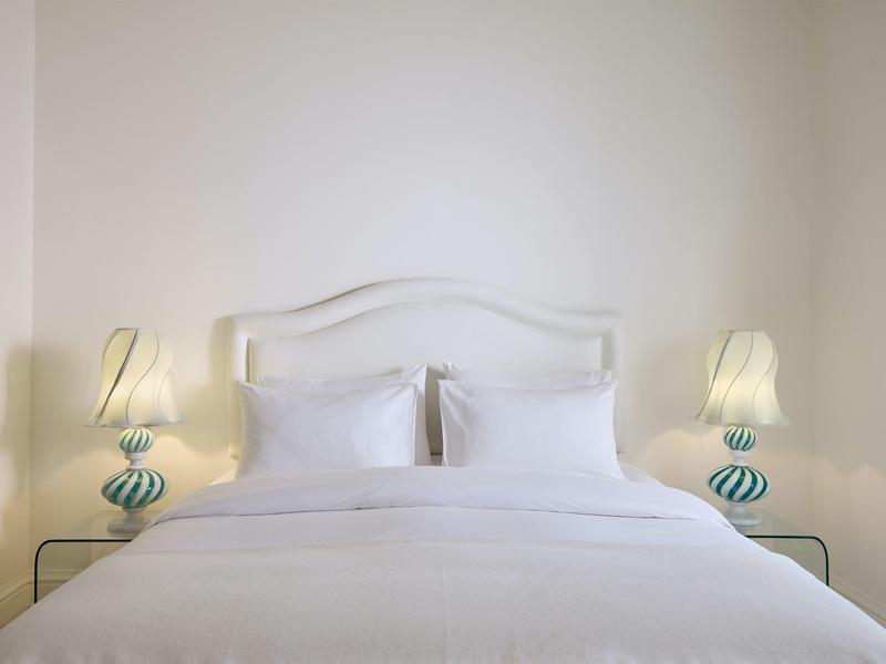 Weißes Bett mit Kissen und zwei Nachttischlampen mit blau-weißen gestreiften Basen vor weißer Wand