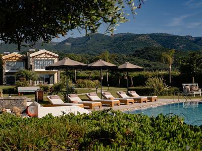 Poolbereich mit Liegestühlen und Sonnenschirmen vor einem Hotelgebäude in den Bergen.