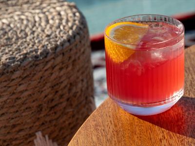 Ein roter Cocktail mit Eis und einer Orangenscheibe auf einem Holztisch neben einem gewebten Stuhl.