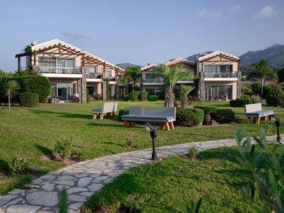 Zwei moderne Villen mit roten Dächern umgeben von grünem Garten und Bergen im Hintergrund.