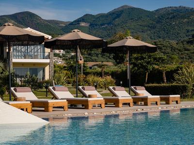 Sechs Sonnenliegen mit Sonnenschirmen an einem Pool vor Bergen und Häusern bei klarem Himmel.