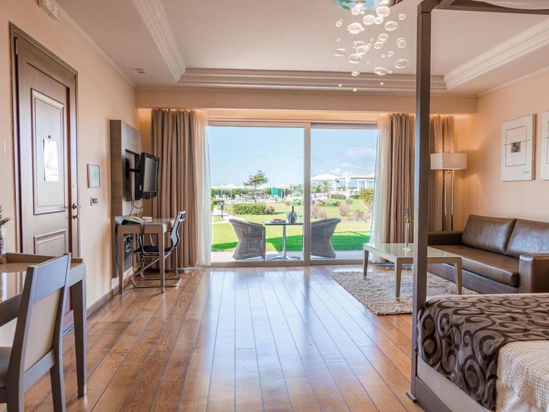 Helles Hotelzimmer mit Holzfußboden, Sitzecke und großem Balkon mit Blick auf grünen Rasen.