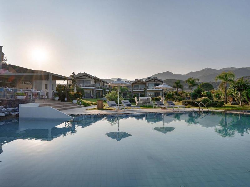 Großer Pool mit Sonnenschirmen und Liegestühlen, umgeben von Gebäuden und Bergen bei Sonnenuntergang.