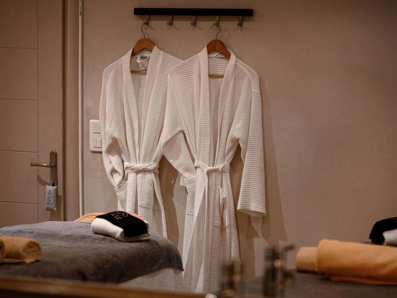 Zwei weiße Bademäntel hängen an der Wand über einem Massagetisch mit Handtüchern.