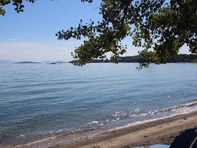 Klares Meer mit sanften Wellen, Sandstrand und grünen Blättern im Vordergrund unter blauem Himmel.
