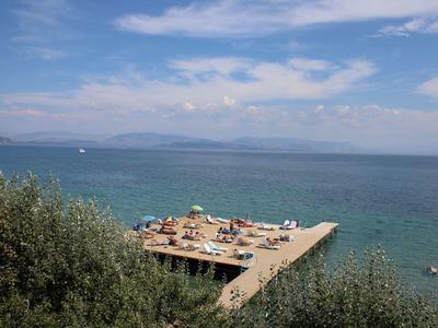 Meeresblick mit blauem Himmel, Bootssteg und Personen auf Sonnenschirmen am Ufer.