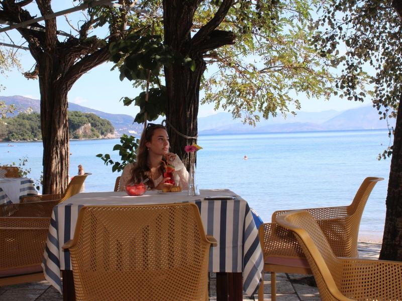 Frau sitzt an mit gestreifter Decke bedecktem Tisch unter Bäumen am Seeufer mit Bergen im Hintergrund.
