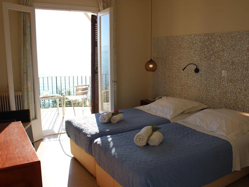 Schlichtes Schlafzimmer mit Doppelbett, blauer Bettdecke, offener Balkontür und Meerblick im Hintergrund.