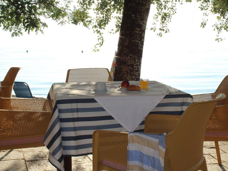 Tisch mit gestreifter Tischdecke, vier Stühle, unter Baum am Wasser bei Sonnenschein.