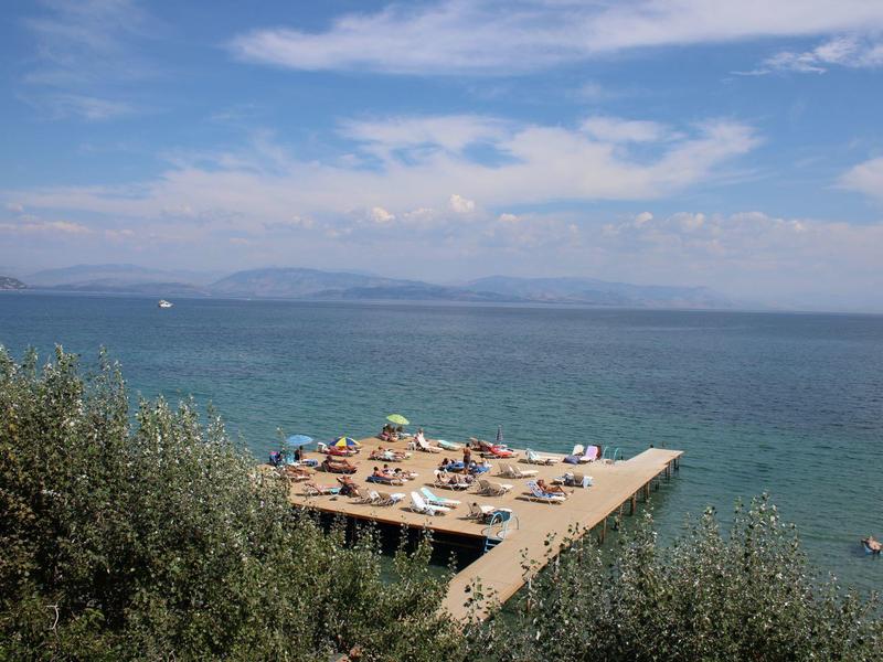 Meeresblick mit blauem Himmel, Bootssteg und Personen auf Sonnenschirmen am Ufer.