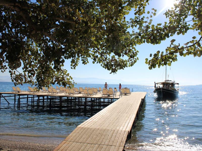 Ein Steg führt über das Wasser zu einem Boot, umgeben von grünem Laub und klarem Himmel.