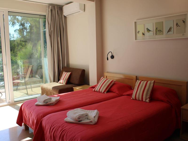 Gemütliches Schlafzimmer mit zwei Betten, roten Decken, Rollos und Blick auf eine Terrasse.