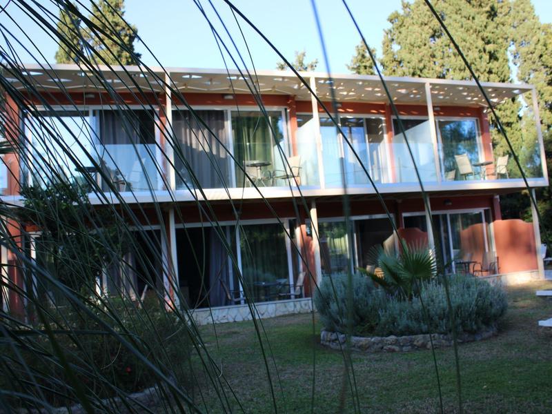 Zweistöckiges Haus mit roter Fassade, großem Garten und Bäumen im Hintergrund bei Sonnenschein.