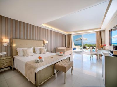 Bright hotel room with two beds, seating area, and glass doors opening to a balcony with sea view.