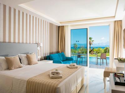 Helles Hotelzimmer mit Doppelbett, blauem Sessel und Blick auf Meer und Terrasse.