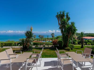 Blick von einer Terrasse mit Stühlen auf einen Garten und das Meer bei klarem Himmel.